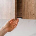 Hand opening a light brown kitchen cabinet. White wall in the background.